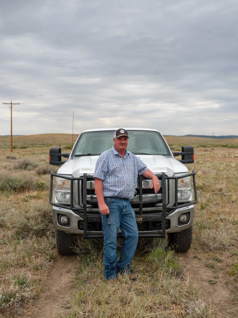 A man in plaid shirt, jeans and a baseball cap stands in front of a white truck in a grassy field in Colorado.
