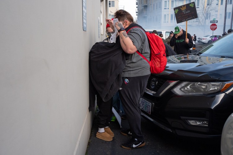 A child stands up against a wall in a parking lot with a sweatshirt draped over them, covering their head and body. A woman stands over the child, holding her hands up protectively. In the background are protesters and a cloud of tear gas in the air.