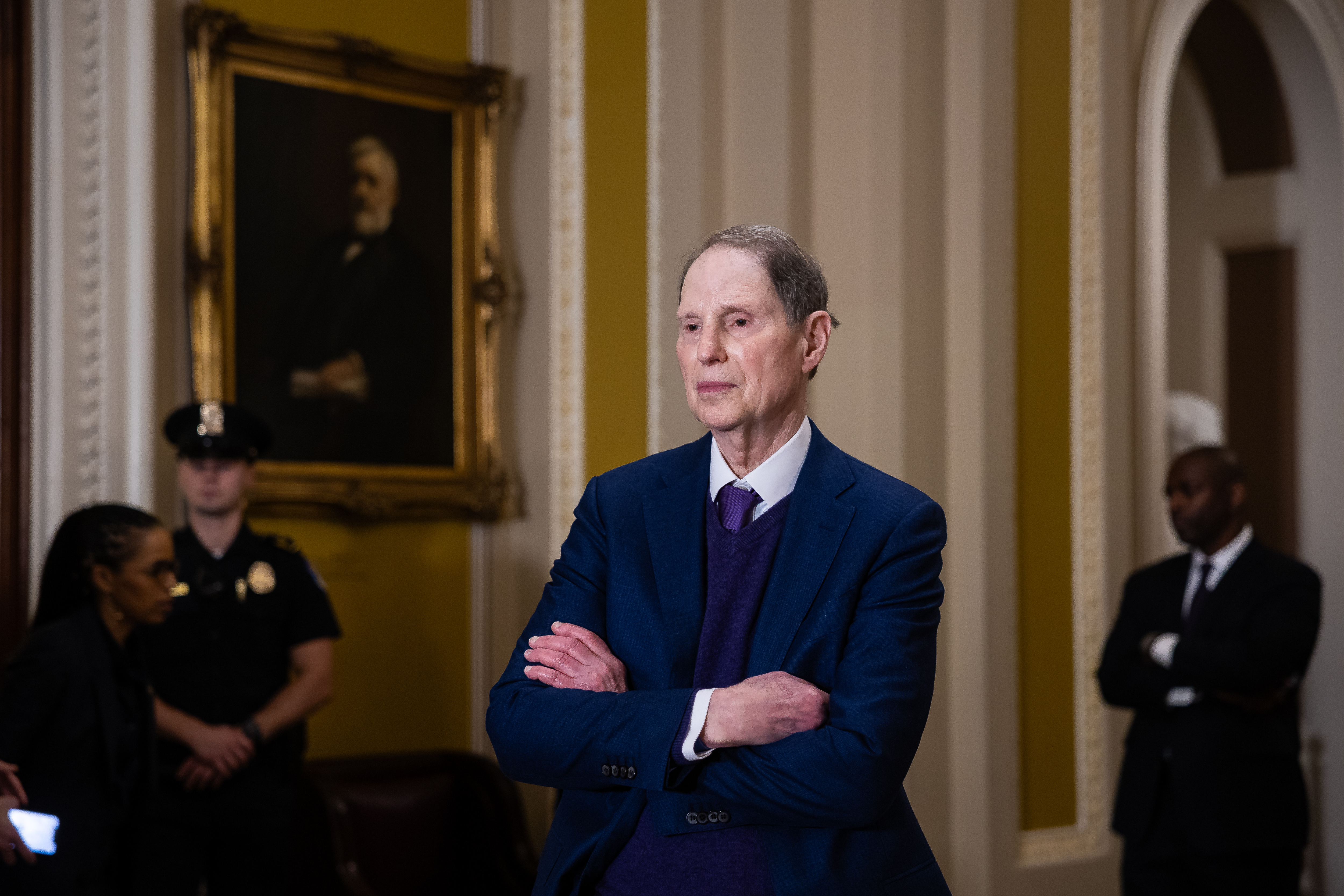 A man with gray hair wears a navy suit and tie and crosses his arms. In the background are three people, including a police officer and a man also crossing his arms wearing a black suit and white shirt. They are all standing in a room with an ornately framed portrait and gold-and-white walls with curved archways.