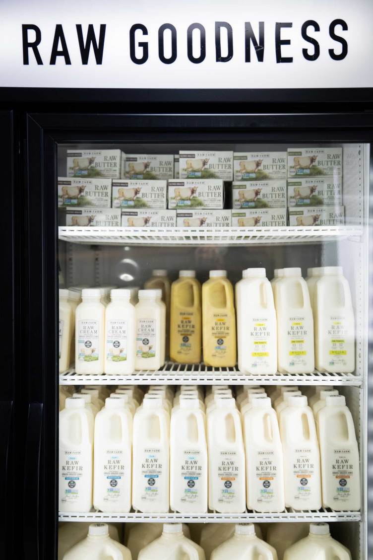 A refrigerator holds multiple plastic containers filled with liquid substances. The labels on the bottles read “raw cream” and “raw kefir.” On the top shelf of the refrigerator are small boxes that read “raw butter.” The refrigerator has text at the top that reads “raw goodness.”