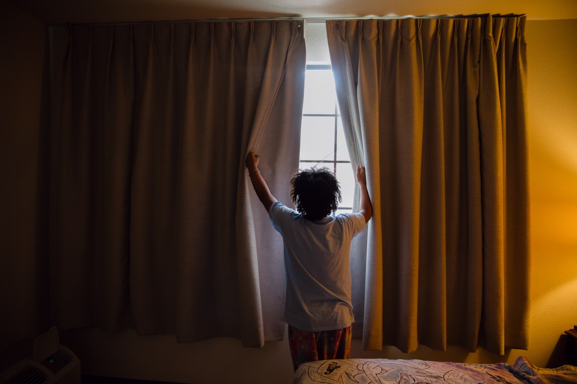 A young child wearing pajamas looks out the window as she opens the curtains to let light into a hotel room.