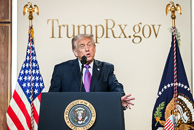 Trump speaks at a lectern in front of a backdrop that says TrumpRx.gov in gold lettering