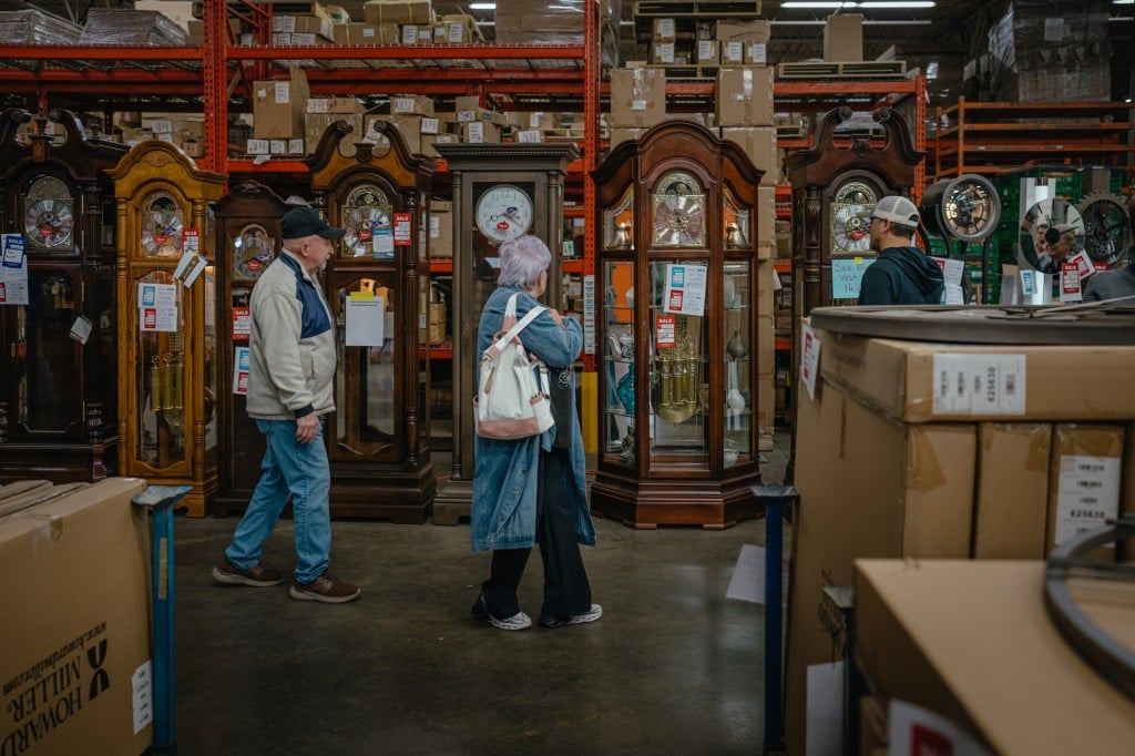 Three people examine a line of grandfather clocks for sale at a warehouse, surrounded by stacks of cardboard boxes and metal shelving.