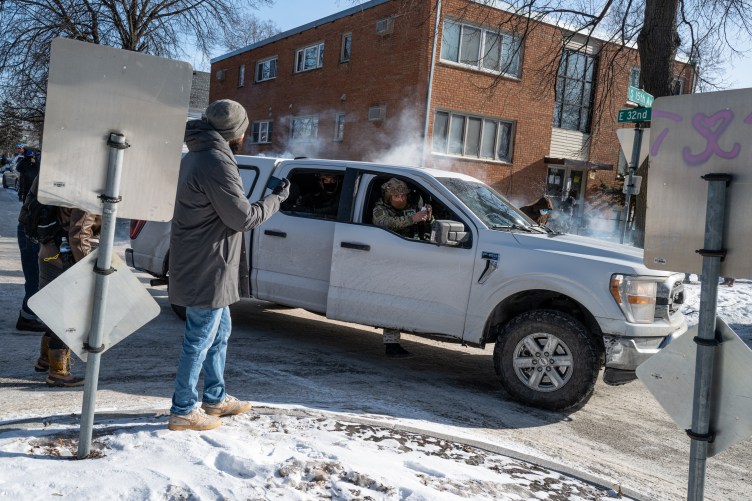 Men in camo wearing helmets and masks  open the doors of a four-door pickup truck while holding weapons and a gas canister. Outside the truck, a few people in street clothes are gathered in a snowy suburban roundabout. One is filming on his phone. 