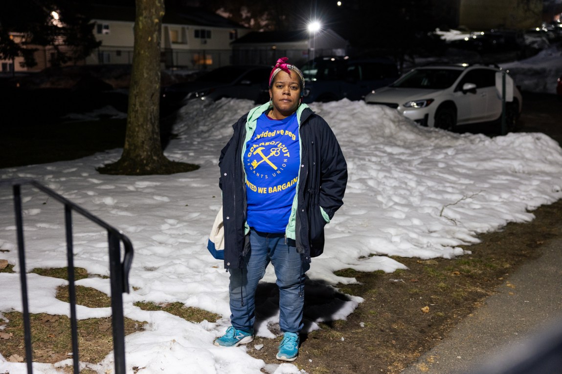 A woman wearing multiple jackets and a shirt that says, “Connecticut Tenants Union: Divided We Beg, United We Bargain,” stands outside at night. Behind her is grass covered in snow, a parking lot and a building with some lights on.