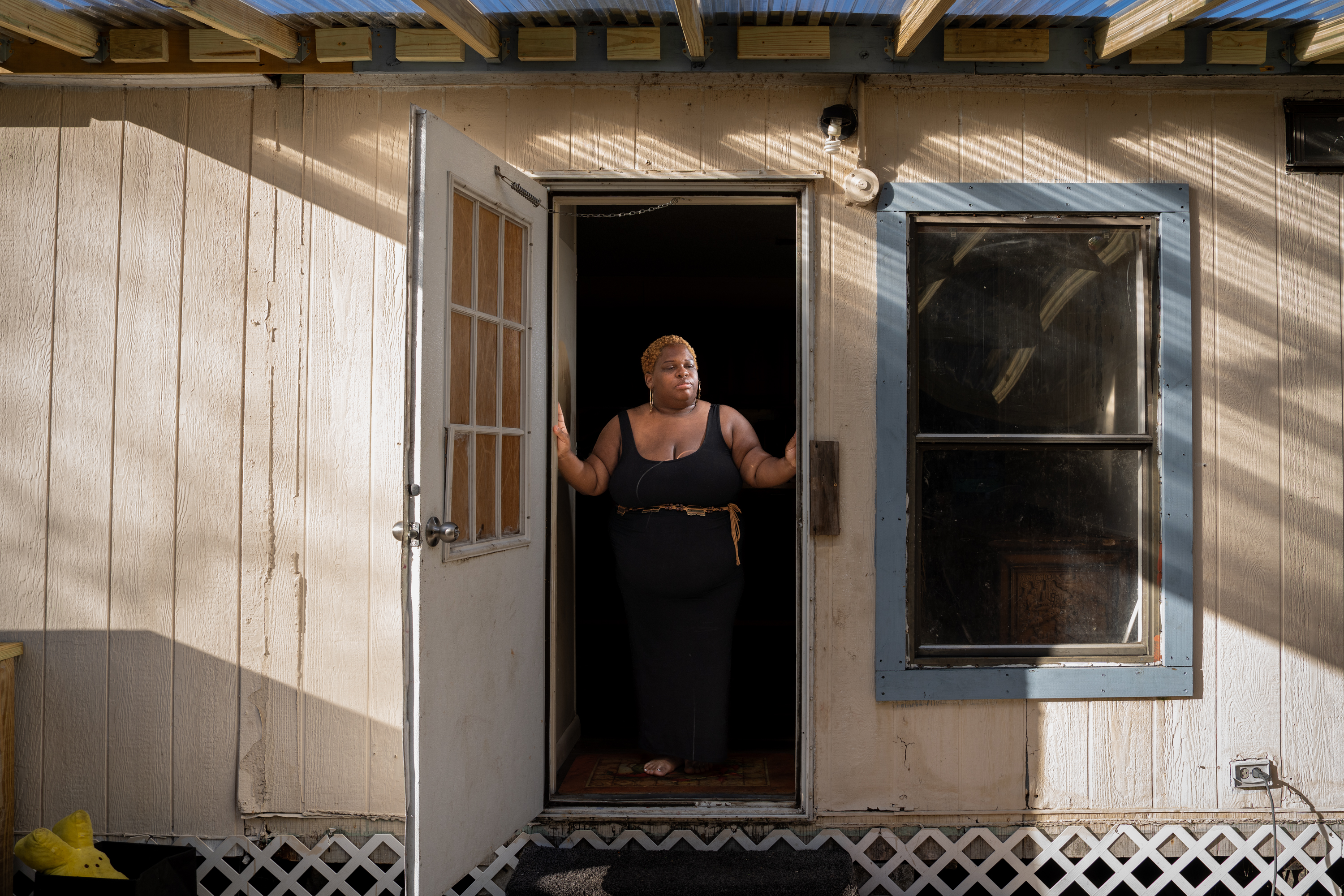 A woman wearing a floor-length black sleeveless dress stands in an open doorway with her hands on the doorframe. A clear porch roof overhead lets light stream in.