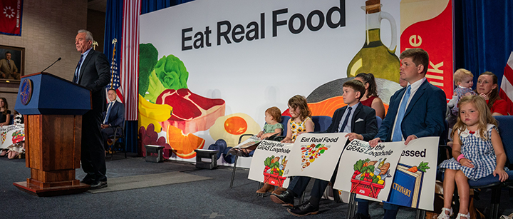 Health Secretary Robert F. Kennedy Jr. stands at a podium in front of a backdrop that says "Eat Real Food" and has images of various meats, vegetables and other foods on it. Behind him and to one side are children holding signs.