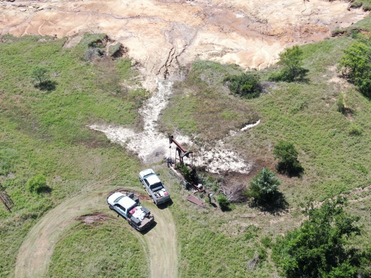 An aerial photo of two trucks parked on grass near an oil well with white salt covering the area downhill from the well.