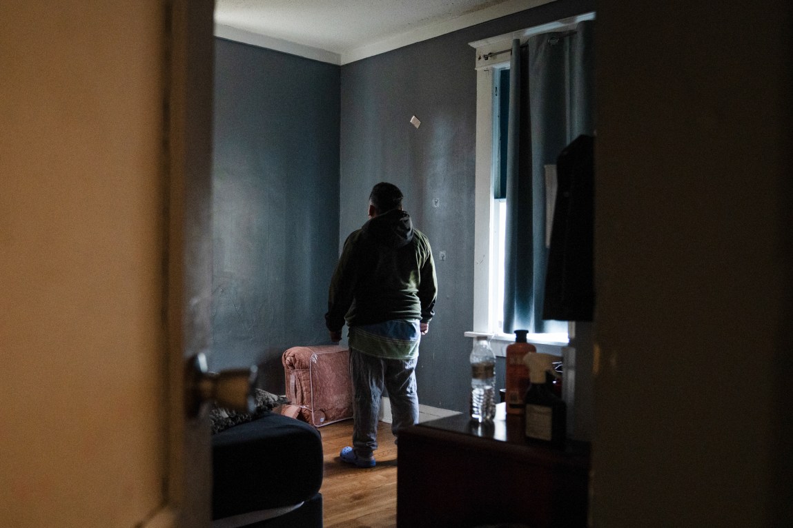 A man stands with his back to the camera in a sparse bedroom. He is framed by an open doorway.