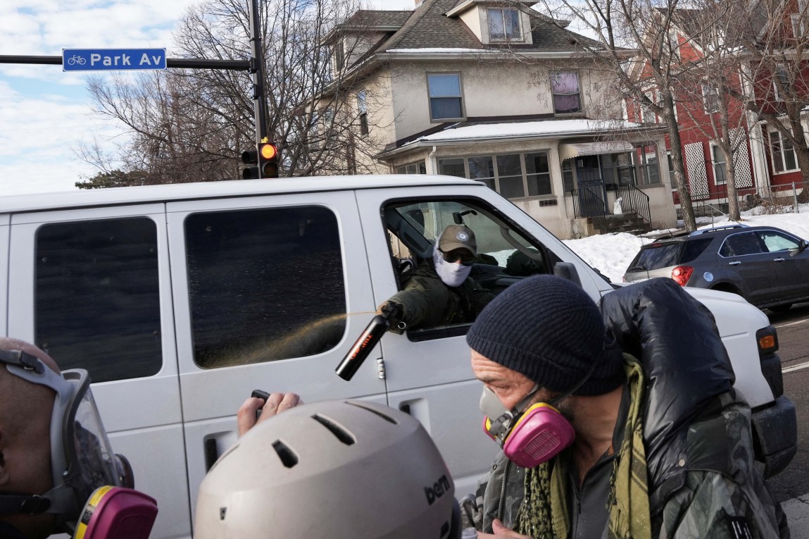 A person with a baseball cap and a balaclava over their face leans out of a white van and sprays an orange substance toward three people wearing air filtration masks and, in one case, a helmet.