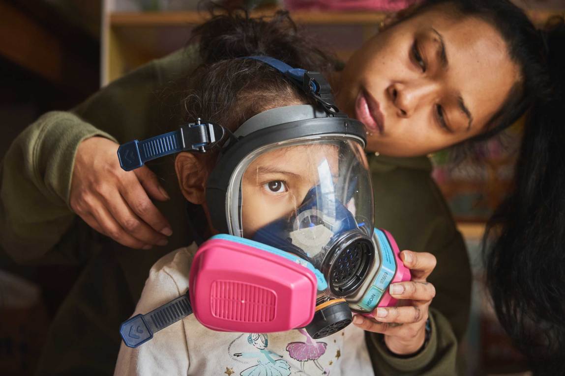 A woman adjusts a large respirator mask with bright pink filters onto a young girl’s face.