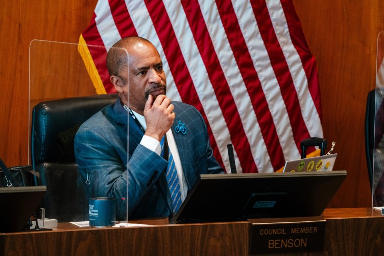 A man wearing a suit holds his chin in his hands while he sits in a government building. In front of him is a placard that reads, “Council Member Benson,” and behind him is an American flag.