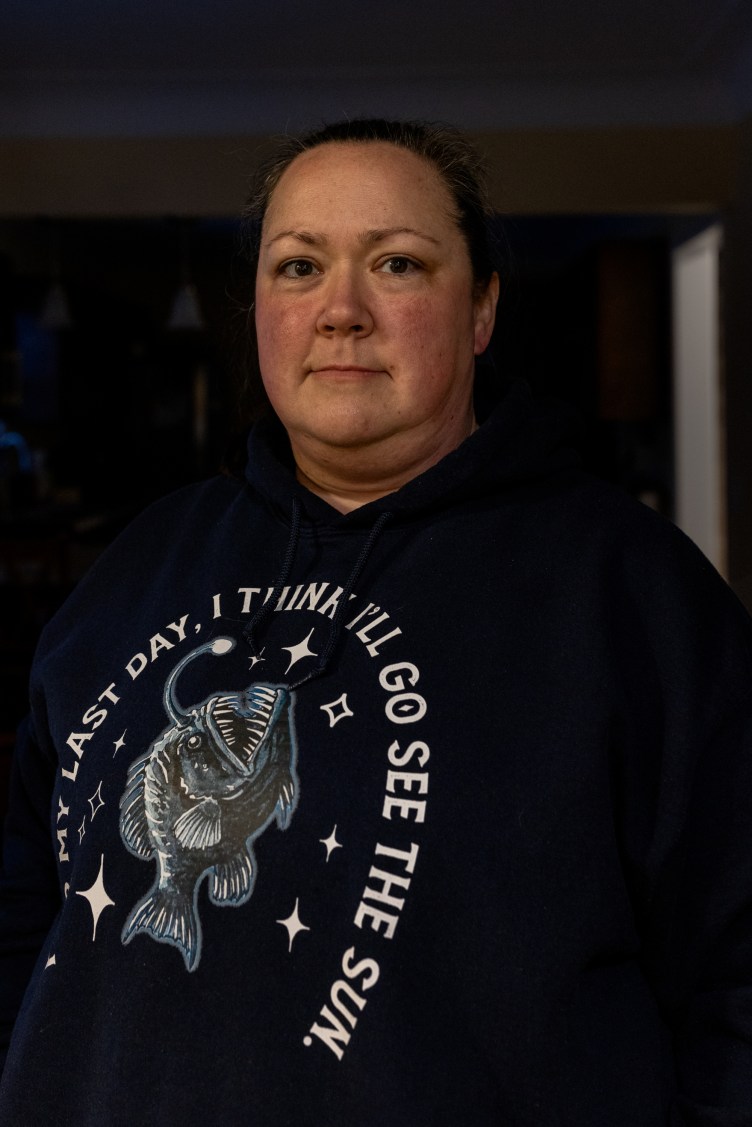 A woman in a dark room looks at the camera. She wears a sweatshirt with an image of an anglerfish and the words “my last day, I think I’ll go see the sun.”