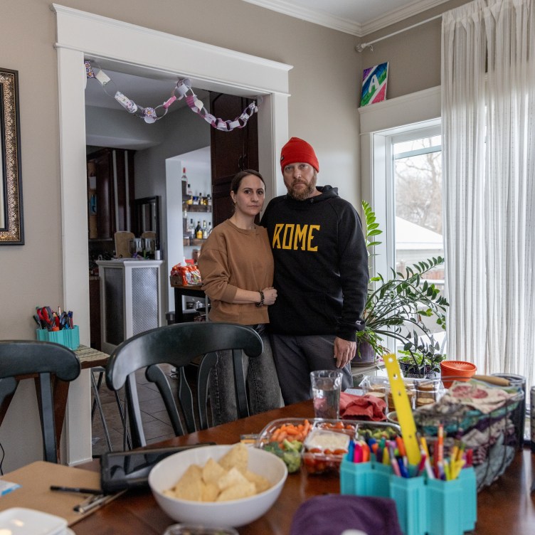 A woman and a man stand together in a dining room. In front of them, a dining room table is covered with snacks, drinks, a pencil holder full of pencils, a laptop and a tablet. On a window next to them, a large sheet of paper is taped up with meeting notes written on it.