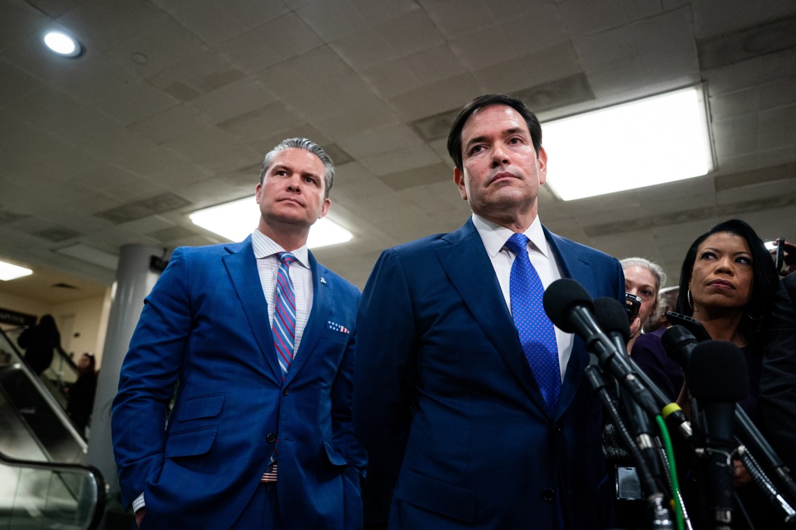 Two men wearing blue suits, with serious expressions, stand in front of news microphones.