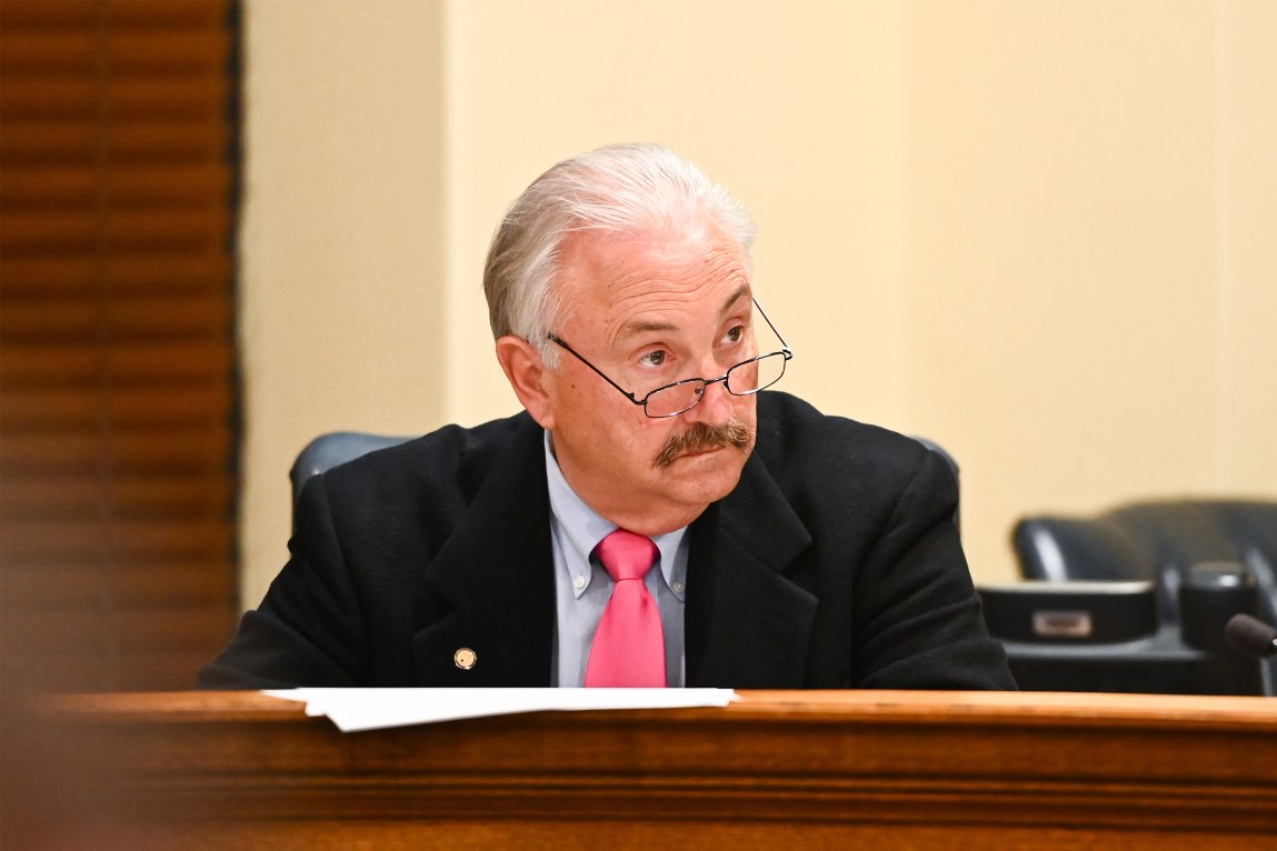 A man with white hair and a black moustache looks off camera. He is wearing a suit and sitting behind a wooden table.