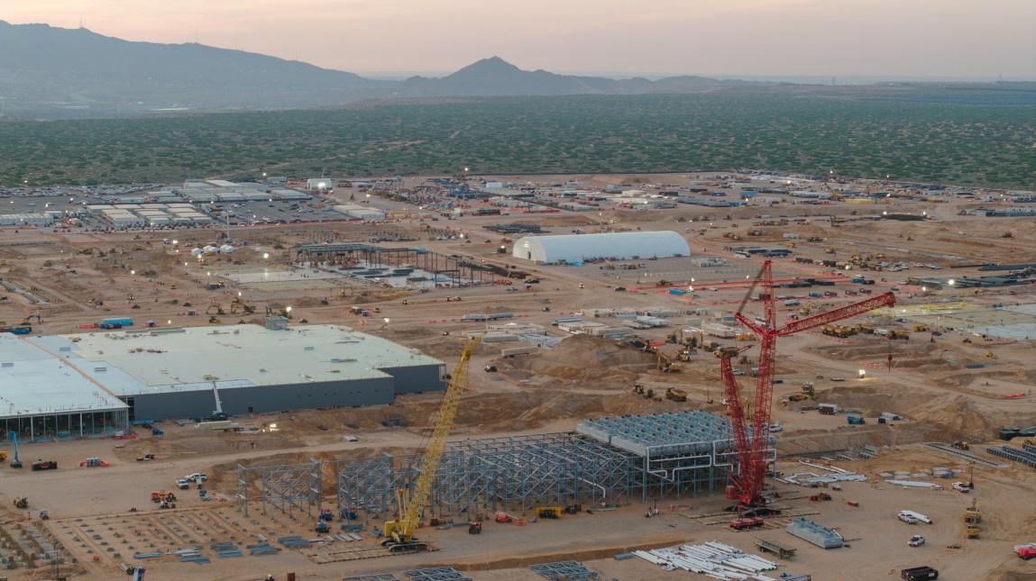 An expansive aerial view reveals a vast construction site in a desert landscape, featuring a large red crane surrounded by neatly arranged building supplies, earthmoving paths and distant mountain ranges.