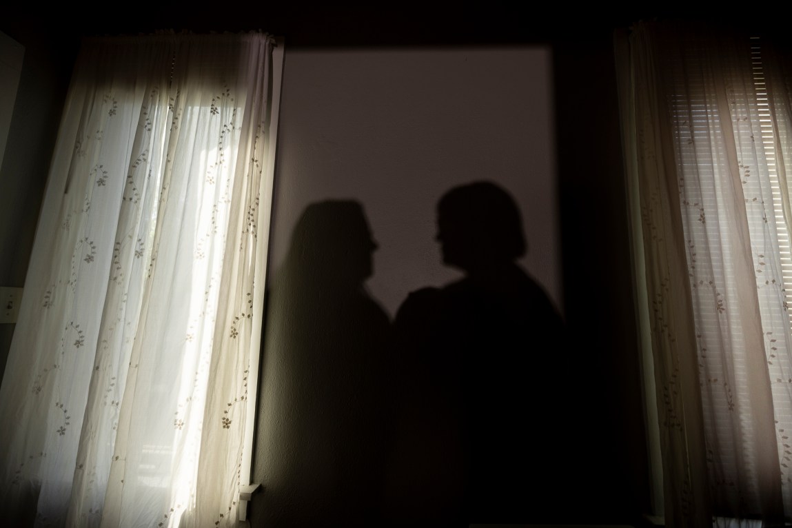 Two women's shadows are reflected on a bedroom wall. On either side of their shadows are curtains with light shining through.