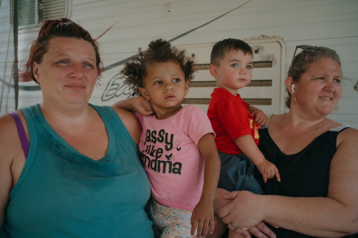 A woman wearing a blue tank top holds a child wearing flower-print pants and a pink T-shirt that says, “Sassy like Grandma.” Another woman wearing a black tank top and earbuds holds a child wearing a red T-shirt and blue pants. The group stands in front of a trailer.