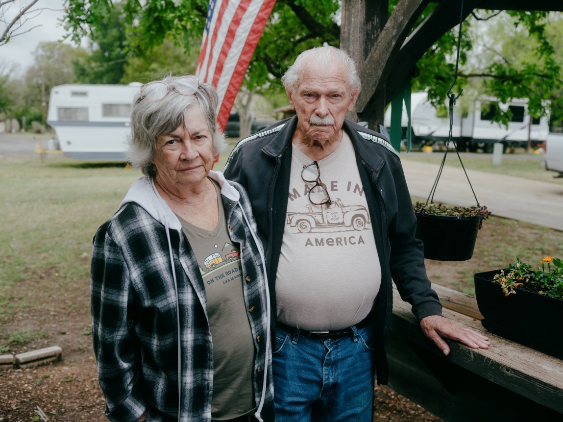 An elderly couple standing outdoors next to a wooden bench, with camping trailers and an American flag visible in the background.
