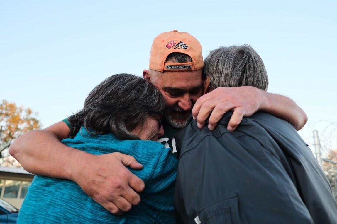 A man wearing a backward baseball cap holds two people in a tight embrace.