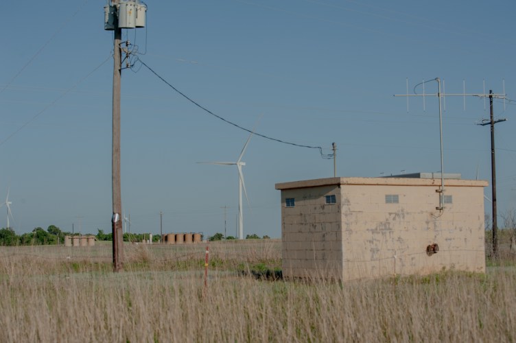 A small beige building with an antenna in a dry, grassy field. Power lines, a wind turbine and a set of brown structures are nearby.