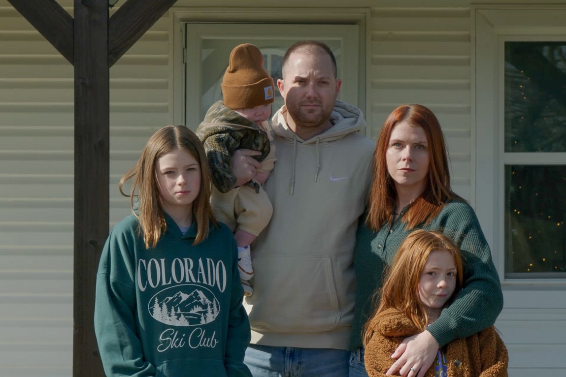 A family of five — a man holding a baby boy, a woman and two girls — stand together outside looking at the camera.