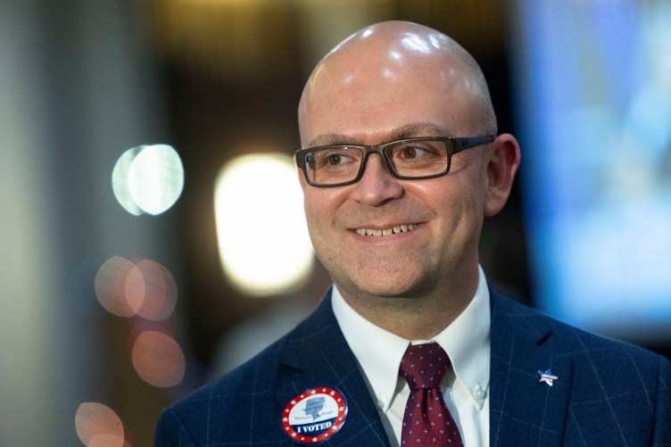 A smiling bald man wearing glasses, a suit and an “I voted” sticker looks off camera in front of an abstract background of blurred lights.