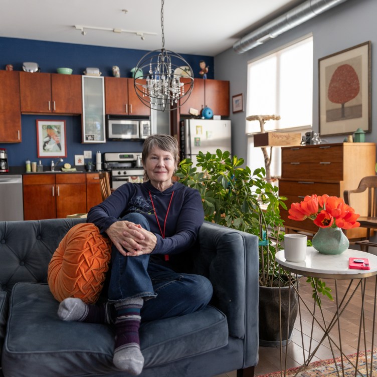 A woman sits with her hands clasped over her leg, looking at the camera. She is on a sofa in the living room of a condo, with several houseplants, paintings on the walls, a full bookshelf and her kitchen in the background.
