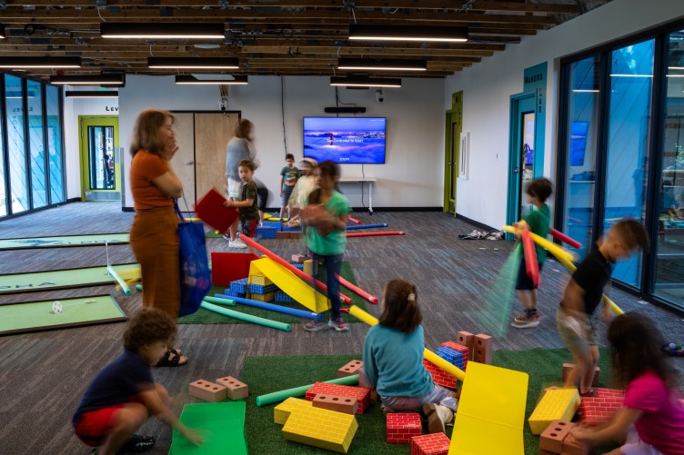 Children play with colorful bricks and other toys in a room with a wood floor and green carpets while two adults supervise.