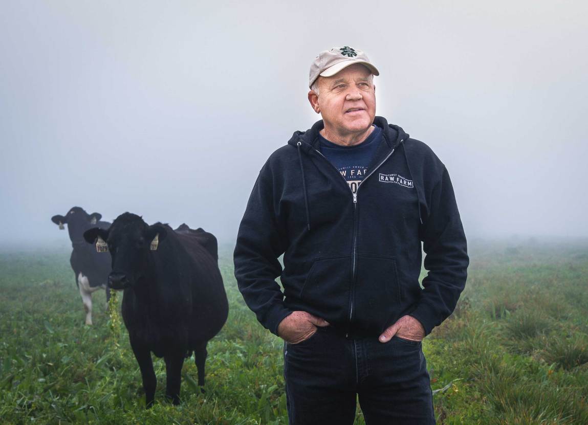 An older man wearing a baseball cap and a black Raw Farm hoodie stands with his hands in his pockets in a foggy, grassy field. Two black cows stand in the background to his right.
