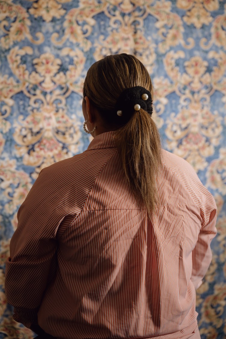 A woman in a striped collared shirt and wearing a scrunchie with pearls on it faces away from the camera. There is an ornate blue wallpaper behind her.