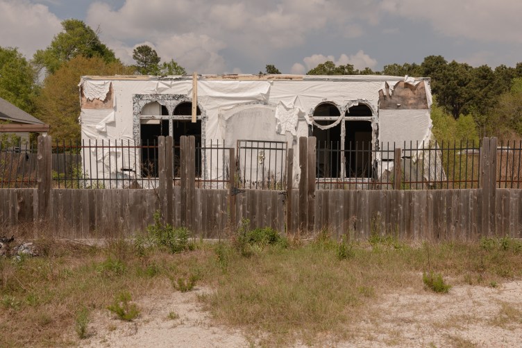 An unfinished house, draped with white plastic construction material, sits behind a deteriorated wooden fence.
