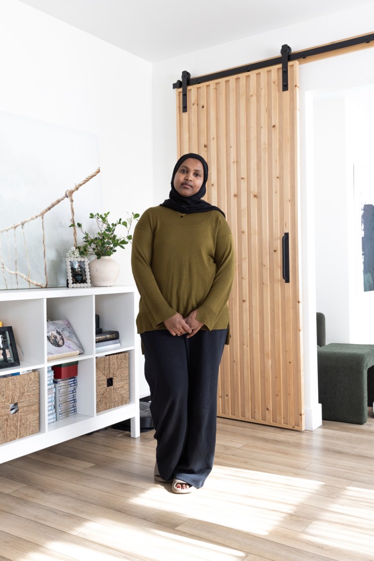 A woman stands looking at the camera in a brightly lit home. Behind her are neatly stacked books and family photos on a shelf.
