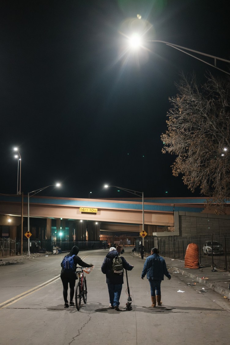 Three individuals, one pushing a bike, another a scooter, walk down a city street at night, illuminated by a street light.