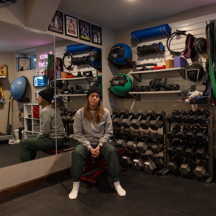 A woman sits in a basement gym, wearing a sweatshirt and athletic pants. She is reflected in a mirror on the wall behind her. There are lines of weights and other gym equipment and a neon sign that says “squeeze your butt.”