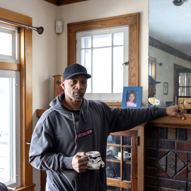 A man stands looking at the camera in his living room, with a mirror and a framed photo of a young girl in the background. He wears a sweatshirt that says “perpetual grind” and holds a Star Wars Stormtrooper coffee mug.