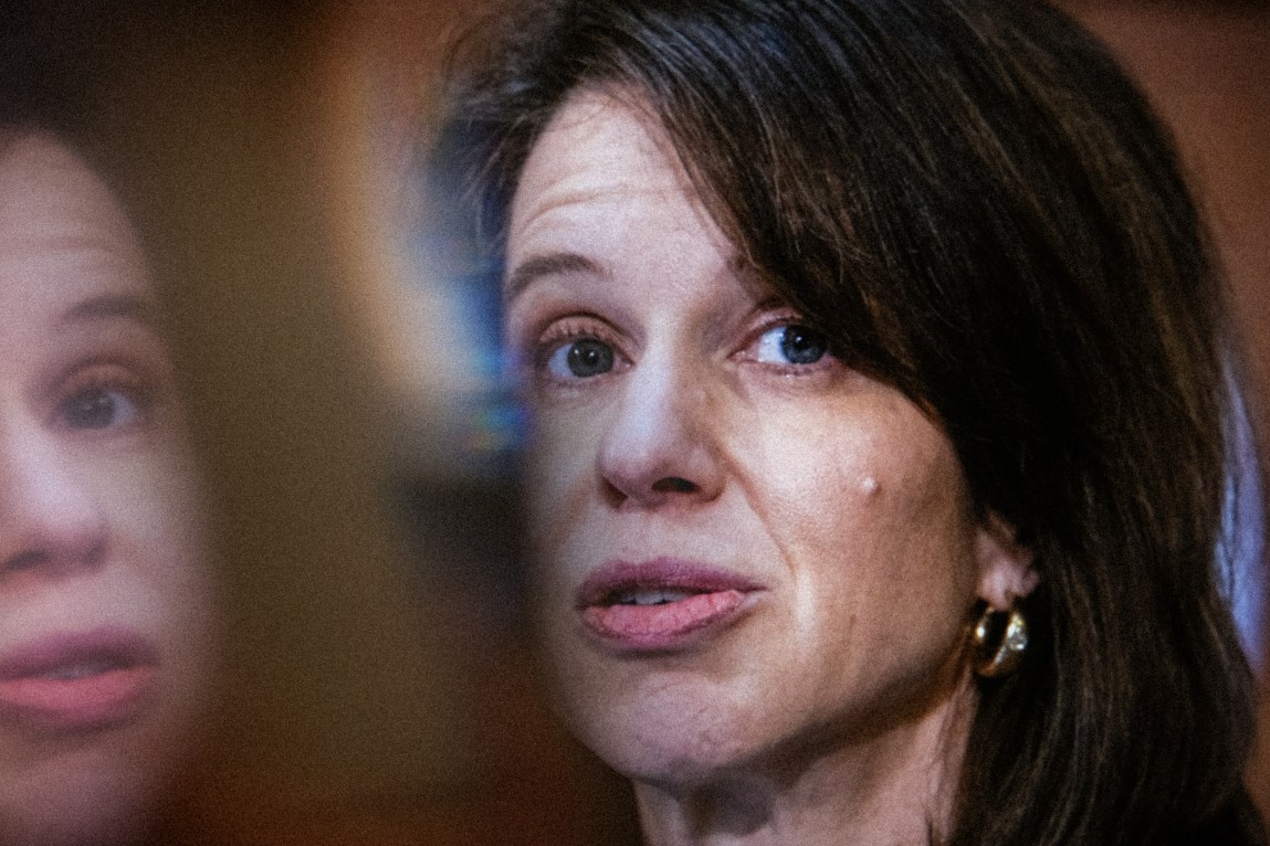 A tightly cropped portrait of a woman in which she appears to be speaking. There is also a blurry reflection of her on the left side of the image.