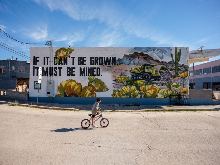 A young boy rides a BMX bike on a road past a large mural depicting mining with the phrase, “If it can’t be grown, it must be mined,” painted on the side of a white building on a sunny day.