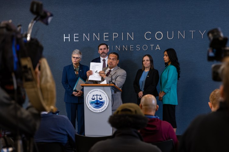 A man wearing a suit holds a piece of paper up while standing behind a podium with a small microphone, flanked by three women and a man, also wearing suits, as reporters and cameras sit before them.