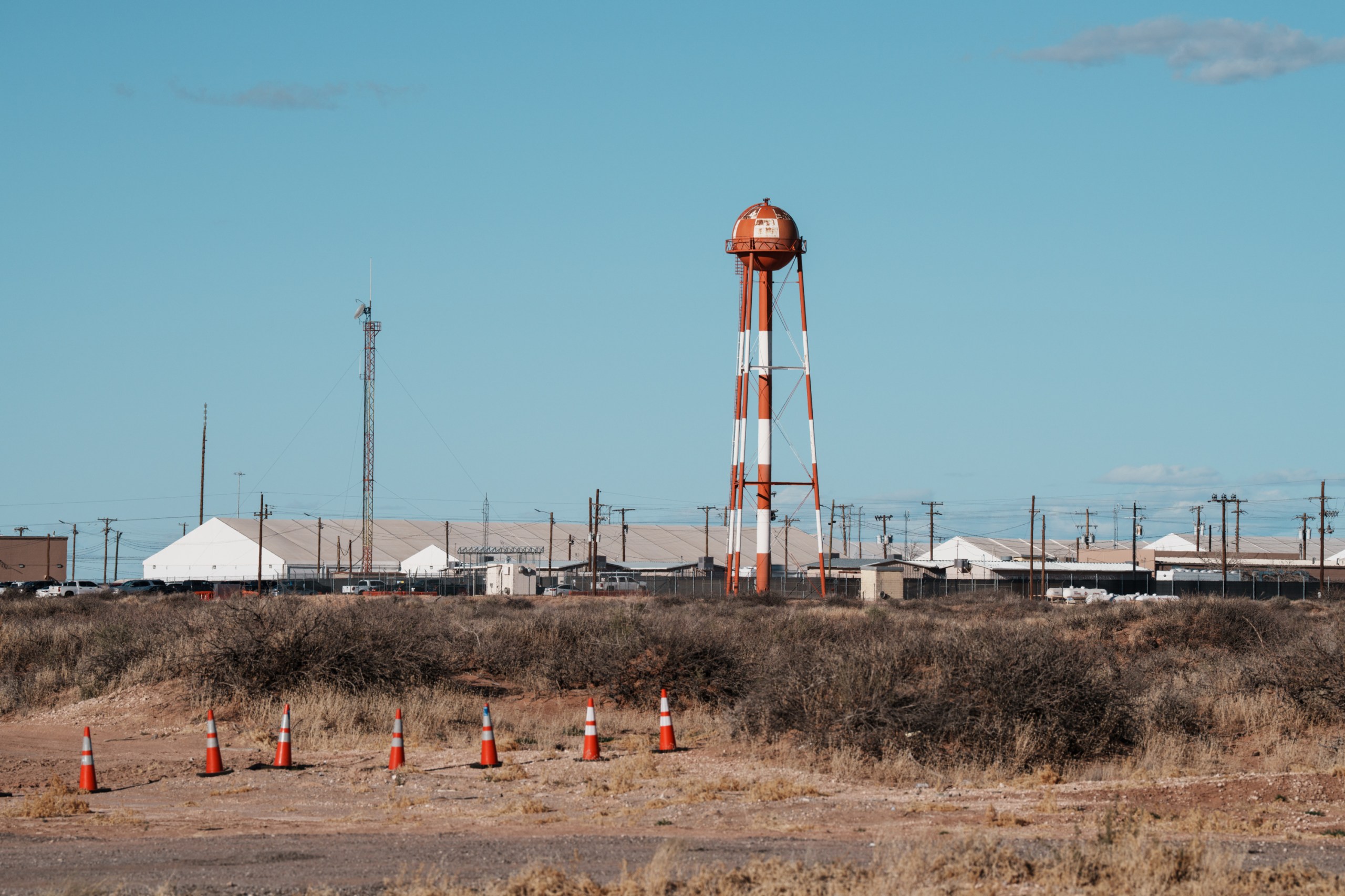 A row of orange traffic cones lines a dry, scrubby dirt field in the foreground. In the background, long, white tent-like buildings and a prominent orange-and-white striped water tower stand under a clear blue sky.