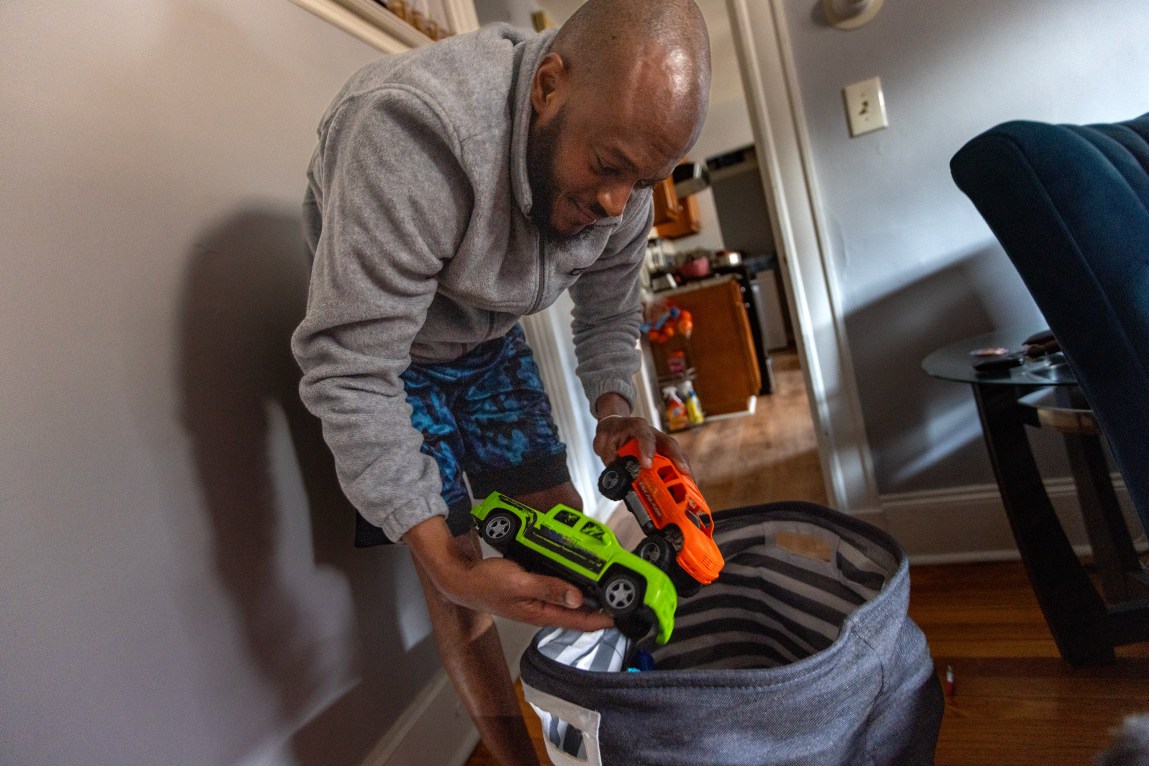 A man leaning over and pulling two trucks out of a basket of toys.