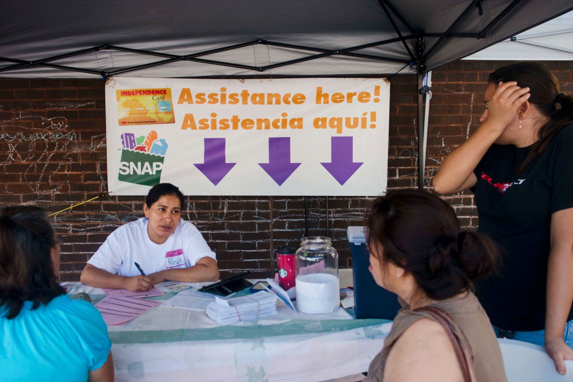 A woman sits at a table with a large sign behind her reading, “Assistance here! Asistencia aquí!” and a SNAP logo. She is writing on a piece of paper while addressing women surrounding the table.