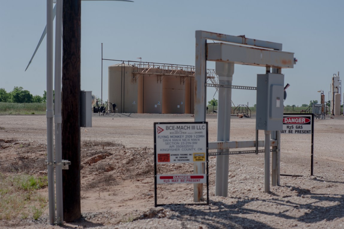 A set of light brown oil disposal well tanks in a dirt lot. Out of focus in the foreground is signage with the words “BCE-MACH III LLC,” “FLYING MONKEY” and “DANGER: H2S GAS MAY BE PRESENT.”