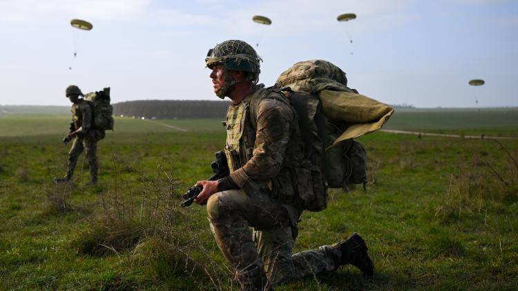 A soldier from 16 Air Assault Brigade kneels, holding an assault rifle, a pack on his back, with parachutes falling in background