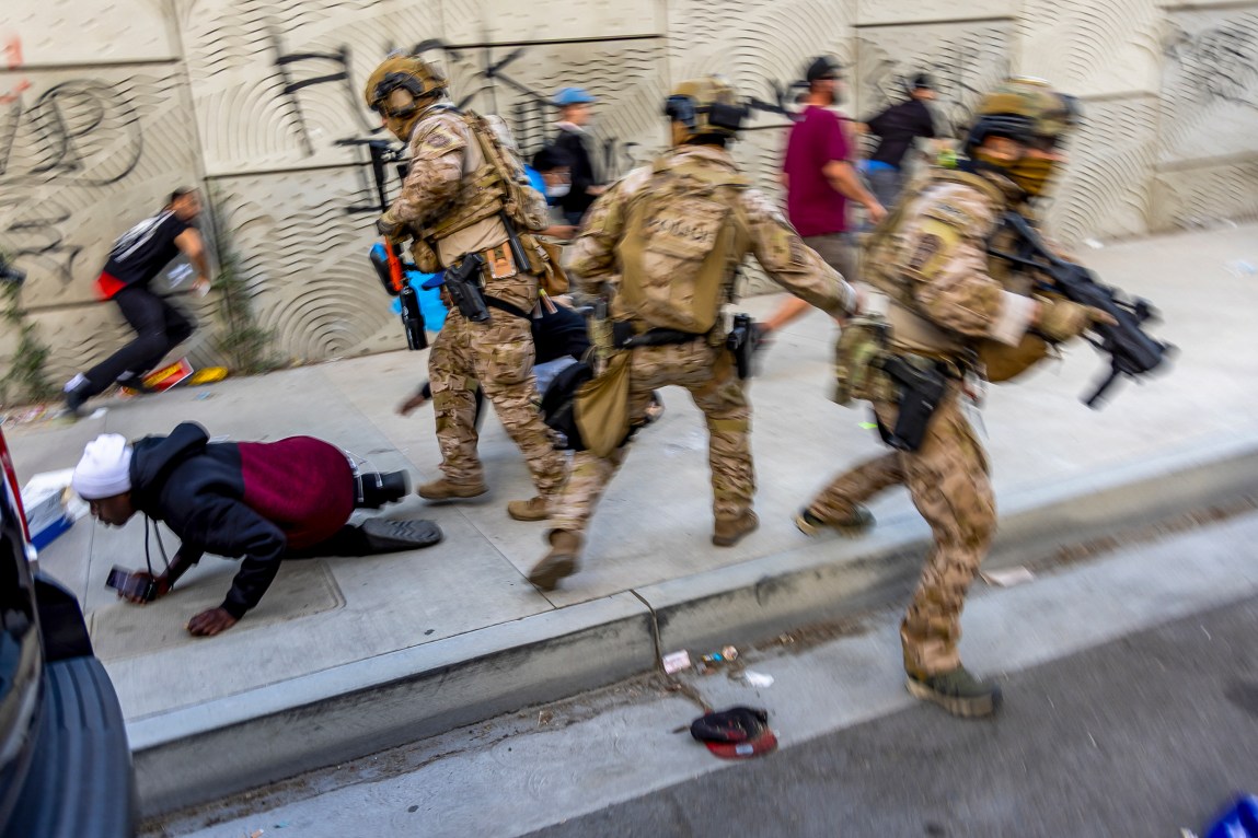 In a hectic scene, three officers with guns, helmets and camouflage gear are a blur of motion on a sidewalk, and all three are moving in different directions. Behind and beside them are people in street clothes, including one person who has fallen on the sidewalk. They are holding a phone and an agent is standing over them.
