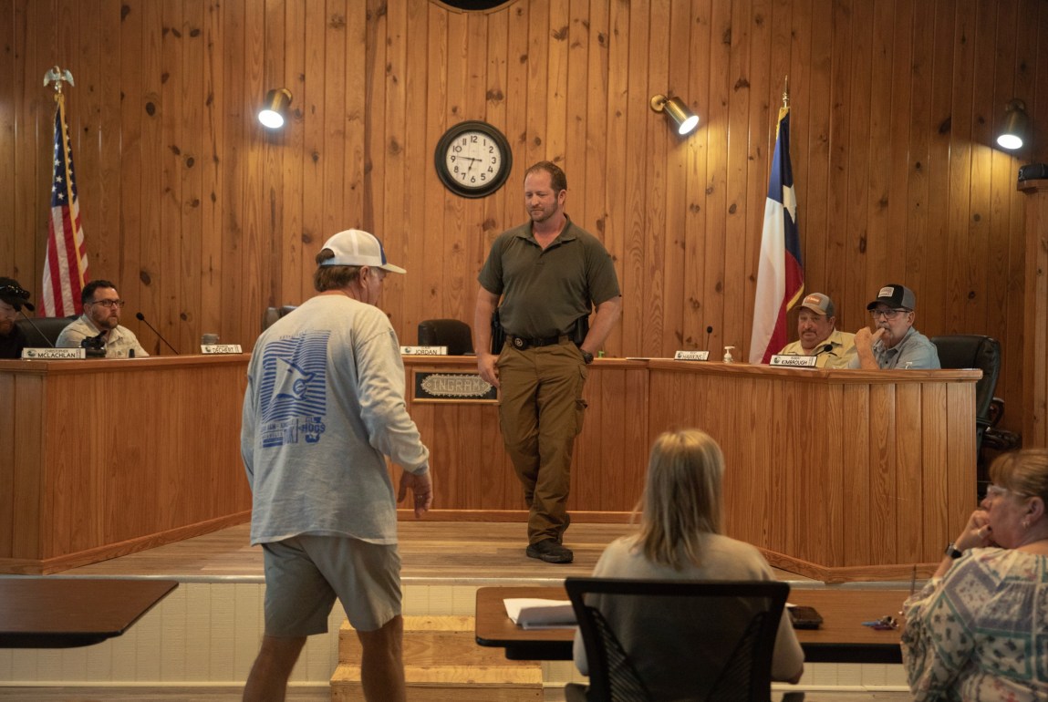 Four men sit at the dais in a wood-paneled room where the Ingram City Council meets, with two other men standing in front and two women seated in the foreground with their backs to the camera.
