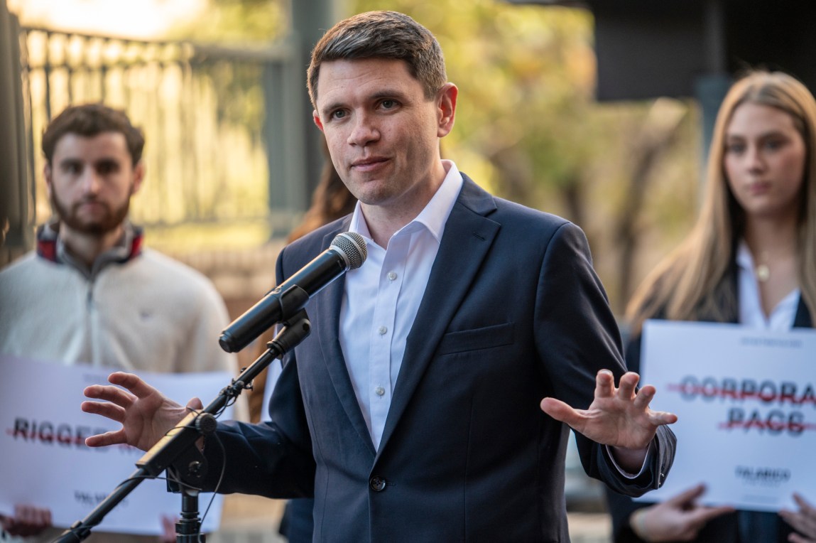 Texas State Rep. James Talarico speaks at a press conference, gesturing with open hands in front of a microphone. Behind him, supporters hold signs with messages opposing corporate PACs and rigged systems.