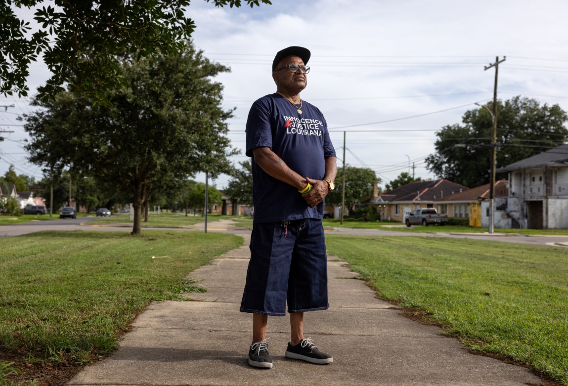 A man wearing denim shorts, a blue T-shirt that says “Innocence & Justice Louisiana,” a baseball cap and glasses holds his hands in front of his torso. He stands on a sidewalk with grass growing on both sides and cars, trees, power lines and houses in the distance. He looks away from the camera.