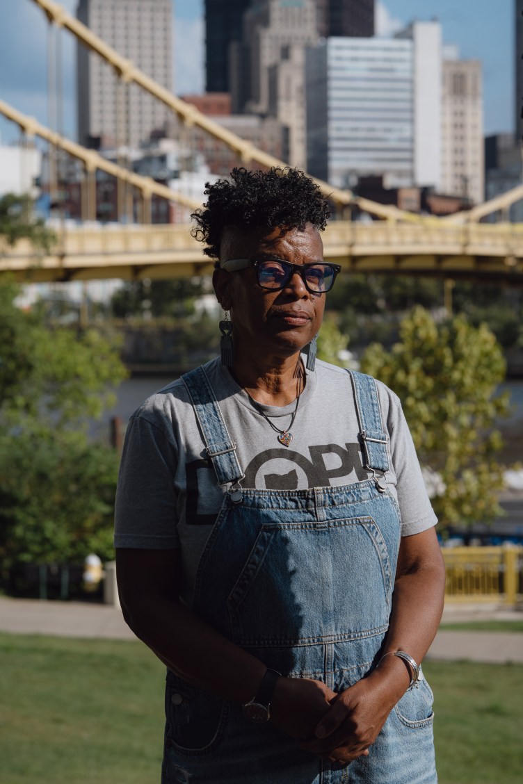 A Black woman wearing denim overalls poses for a portrait in front of the Pittsburgh skyline.